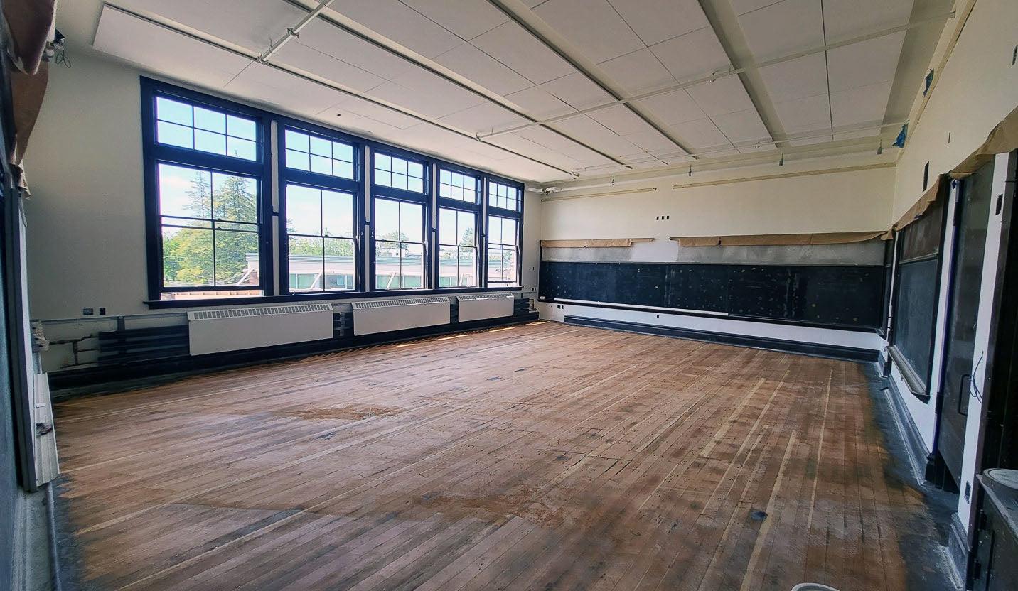 an empty room has blackboards on one wall, windows on another wall with radiators below them, and a wood floor