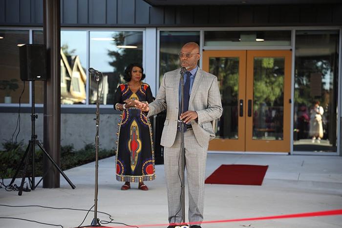 a man wearing a suite stands at a microphone. a woman in a dress stands behind him. both are in front of large glass doors.